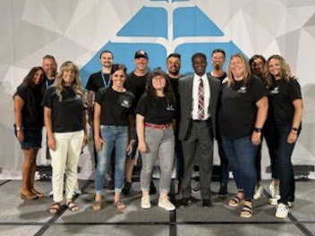 16 staff members on a stage with Dr. Anthony Muhammad at the plc conference smiling for the camera.